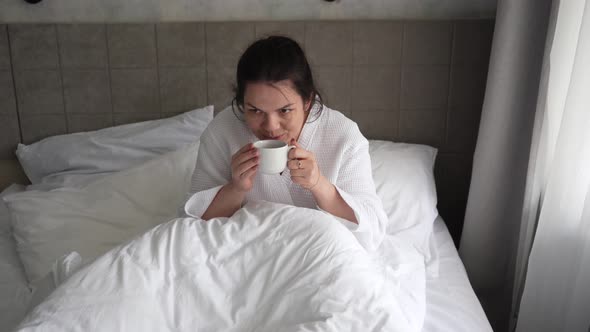 Young Woman in the Morning Lying in Bed and Drinking Coffee Looking at Messages on the Phone