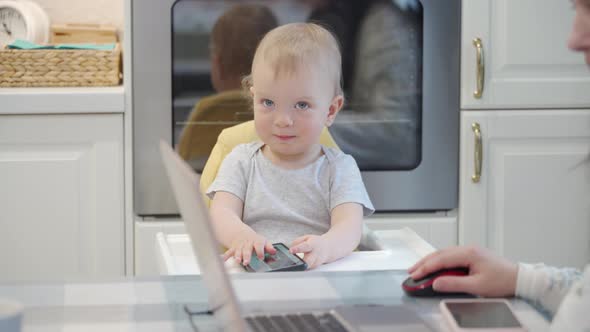 Mother Works on Laptop While Her Toddler Playing with Smartphone One Year Old Baby Boy Sitting in