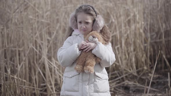 Portrait of Lost Caucasian Little Girl with Teddy Bear Crying Outdoors. Sad Lonely Child Standing at