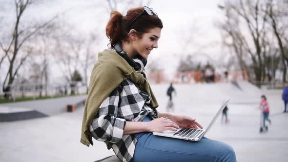 A Young Trendy Brunette is Sitting on a Bench with a Laptop on Her Knees Typing