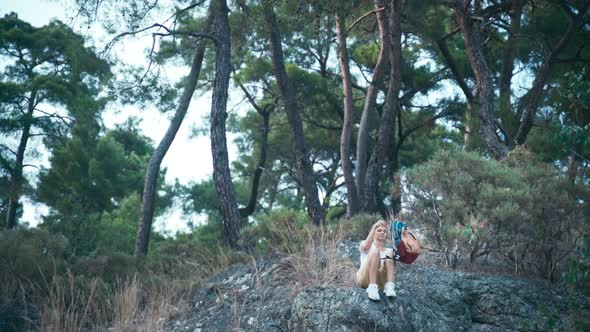 Young Woman with a Backpack Sitting on a Rocky Mountain and Drinking Water