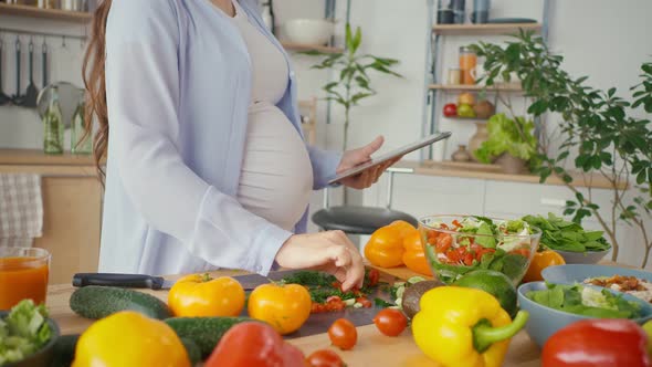 A Pregnant Woman Using a Tablet Prepares Healthy Food in a Bright Kitchen