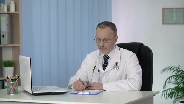 Doctor Writing in Patients Medical Record, Looking Into Camera, Clinic Services