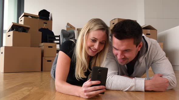 A Smiling Moving Couple Lies on the Floor of an Empty Apartment and Works on a Smartphone