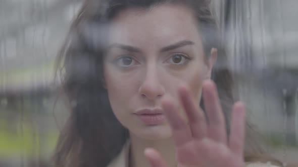 Close-up of Young Caucasian Woman Standing Behind Glass and Touching Window with Hand. Portrait of