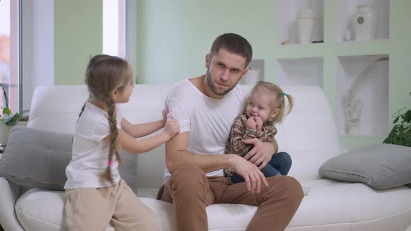 Exhausted Young Single Father Sitting on Couch Looking at Camera As Joyful Girls Tickling Man in