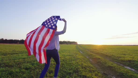 Young Woman Runs with an USA Flag Across a Field at Sunset Rear View