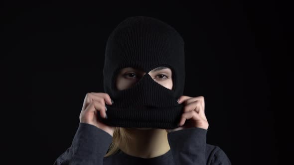 A Young Woman Is Putting on a Balaclava Mask. Bandit on a Black Background Close-up.