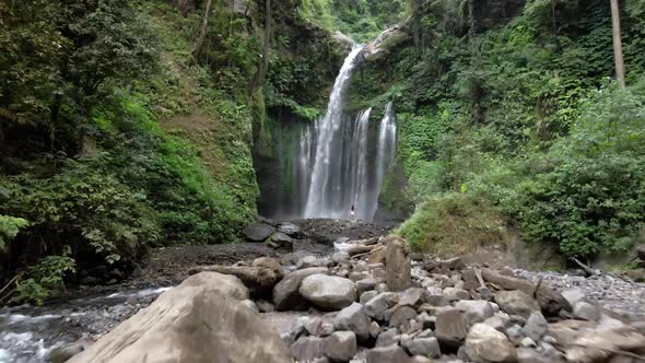 Aerial forward shot between overgrown hills and girl in front of Waterfall,Lombok.