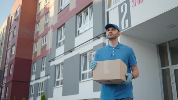 A Delivery Man in a Blue Tshirt and Cap Carries a Cardboard Box in the City Looking Around in Search