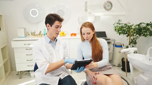 Young Male Dentist and Female Patient Looking at Digital Tablet Discussing Dental Treatment Plan