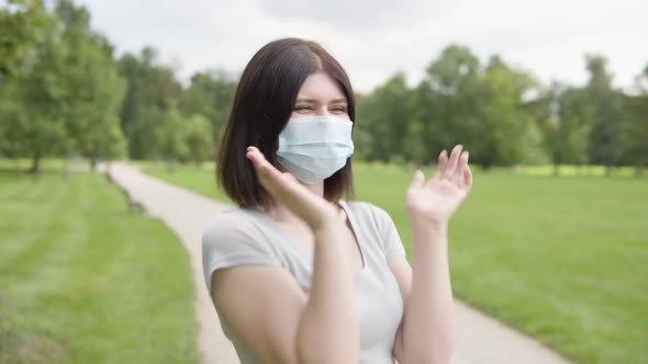 A Young Caucasian Woman in a Face Mask Celebrates on a Pathway in a Park on a Sunny Day