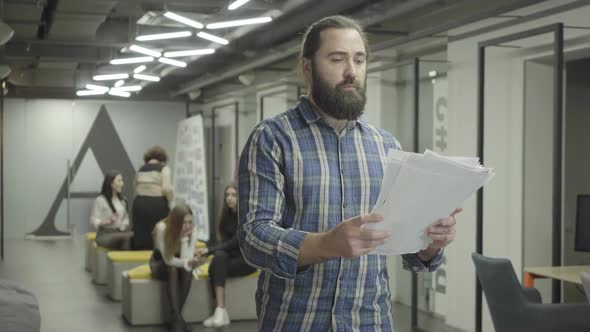 Concentrated Bearded Man Studying Papers Documents Standing in the Office. Young Colleague Riding