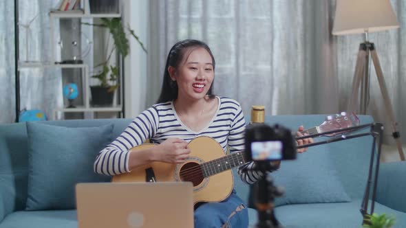 Asian Woman With A Laptop Singing And Playing A Guitar While Live Stream At Home