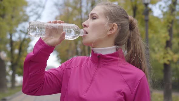 Close-up Portrait of a Young Caucasian Woman in Pink Sportswear Drinking Water and Running Away