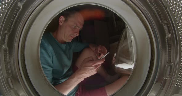 Young Man with Phone in Hands Sits By Washing Machine