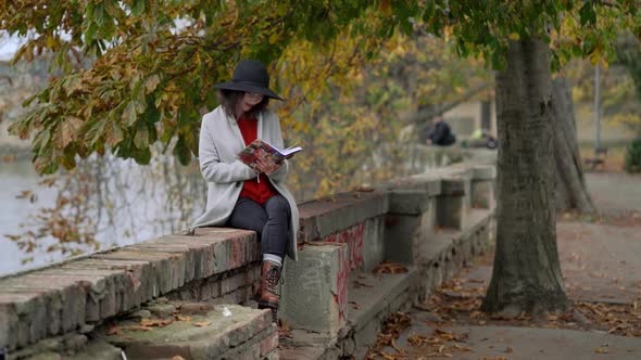 Young Attractive Stylish Woman Reading Book in Park By the River at Autumn Day