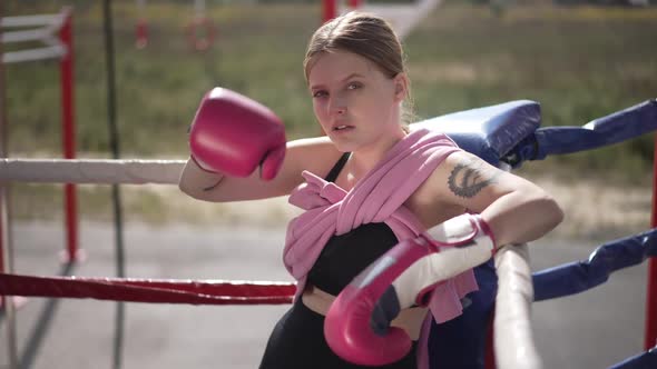 Serious Confident Woman in Boxing Gloves Rubbing Forehead Looking at Camera Sitting Outdoors on