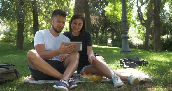 Serious Young Couple or Friends Using Digital Tablet in Park. Man Woman Lying on Grass and