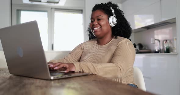 Happy african girl sitting inside kitchen at home using laptop computer while wearing headphones