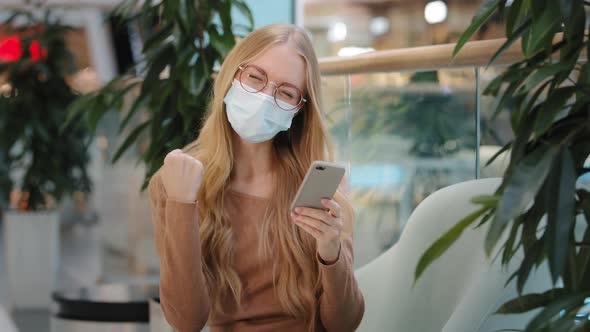 Happy Young Girl in Medical Mask Sit in Mall Got Sms with Good News on Smartphone Joyfully Dancing
