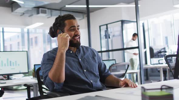 Mixed race businessman sitting at desk looking at computer screen, using smart phone and smiling