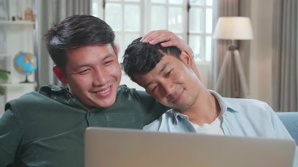 Asian Gentle Gay Couple Using Laptop Computer While Sitting On A Couch In Apartment