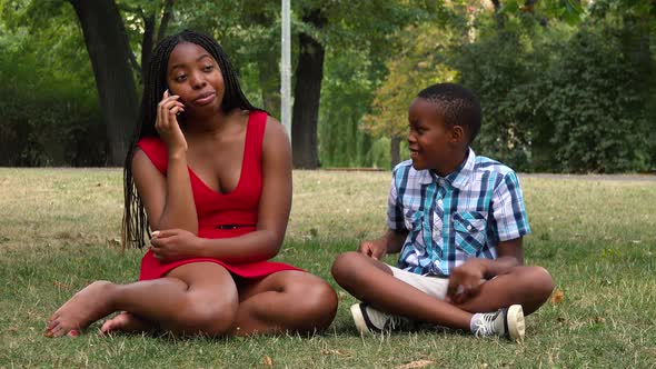 A Young Black Mother and Her Son Sit on Grass in a Park, the Son Tries To Attract the Attention