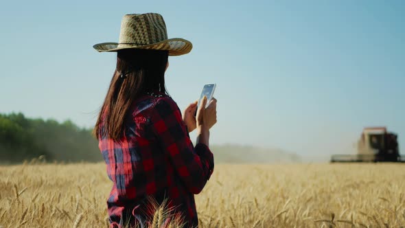 Young Woman Farmer in Hat Standing with a Harvest Against the Background of a Working Combine