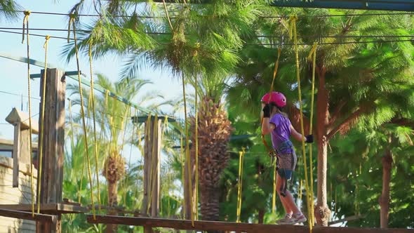 Frightened but Brave Kid Girl in Helmet Tshirt and Shorts Climbs in Rope Park