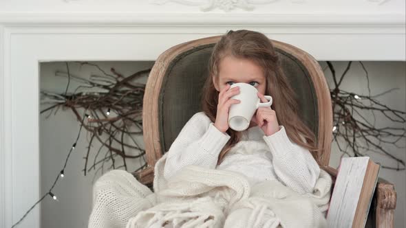 Little Girl Sitting in a Cozy Chair Wrapped in a Blanket Drinking Tea Near Christmas Fireplace