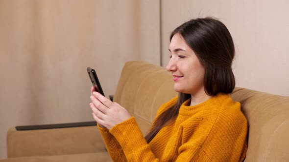 Brunette in Pullover Sits on Sofa and Types on Smartphone