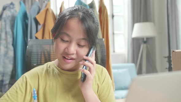 Asian Young Girl Online Seller Talk On Mobile Phone And Take Note For Selling Clothes At Home