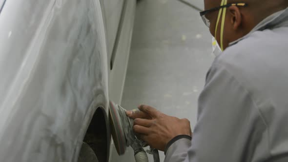 African American male car mechanic wearing a face mask and polishing a side of a car with a grinder