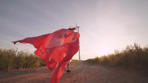 Two Girls with Pieces of Red Cloth Run to the Wind Generator in the Field