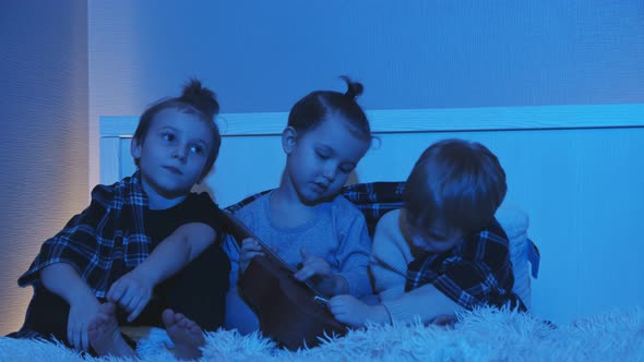 Three Little Boys Sit on Bed and Play Guitar in Room at Night