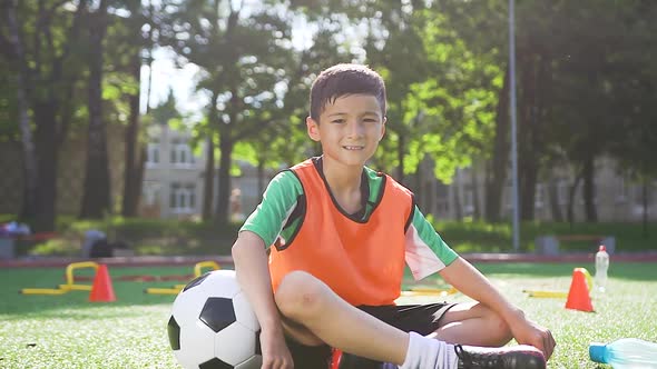 Asian Teen Boy in Orange Vest Sitting on the Artificail Turf with Football Ball