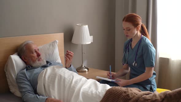 Professional Female Doctor Talking to Senior Adult Male Patient Lying on Bed at Home and Checking