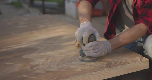 A Man in a Respirator Goggles Polishes a Parquet Board with an Orbital Sander