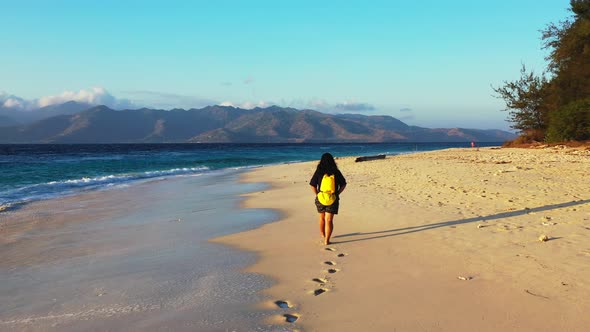 Girl with yellow backpack walking barefoot around exotic beach washed by sea waves at sunrise with v