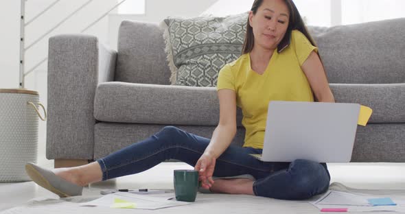 Focused asian woman sitting on floor, drinking coffee and working remotely from home with laptop