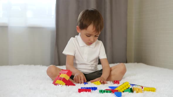 A Happy Child Sits on a Bed in a Room and Builds From Colored Blocks, Playing and Imagining