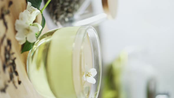 Glass Cup Of Green Tea With Jasmine On Bamboo Table With Sprig Of Jasmine. Hand Puts Fresh Jasmine