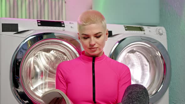Punk Woman with Short White Hair and Pink Jumpsuit Sits on Floor Near Washing Machines and Read