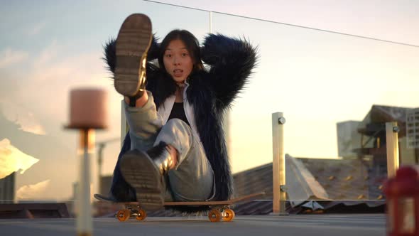 Front View Joyful Young Asian Woman Posing on Urban Roof at Sunset Sitting on Skateboard