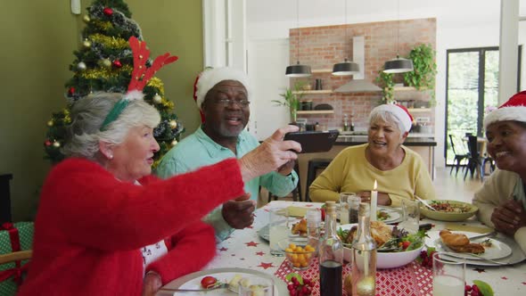 Diverse senior friends in santa hats looking at smartphone and laughing at christmas dinner table