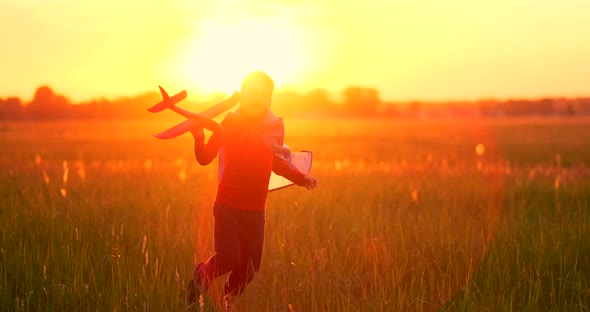 The Boy Runs Across the Field with a Plane in His Hands at Sunset