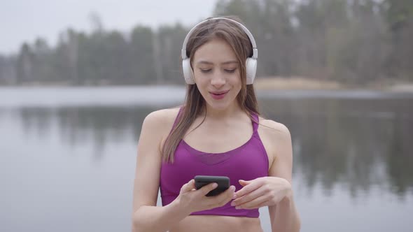 Portrait of Cute Young Woman in Headphones and Sportswear Dancing on Lake Shore and Singing