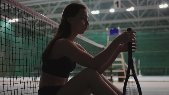 Young Pretty Female Tennis Player is Sitting on Floor on Tennis Court After Match or Training