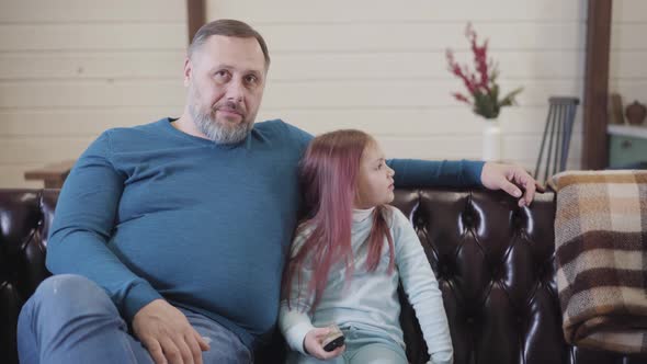 Charming Cheerful Little Girl Sitting with Father on Couch Watching TV and Calling for Mother To